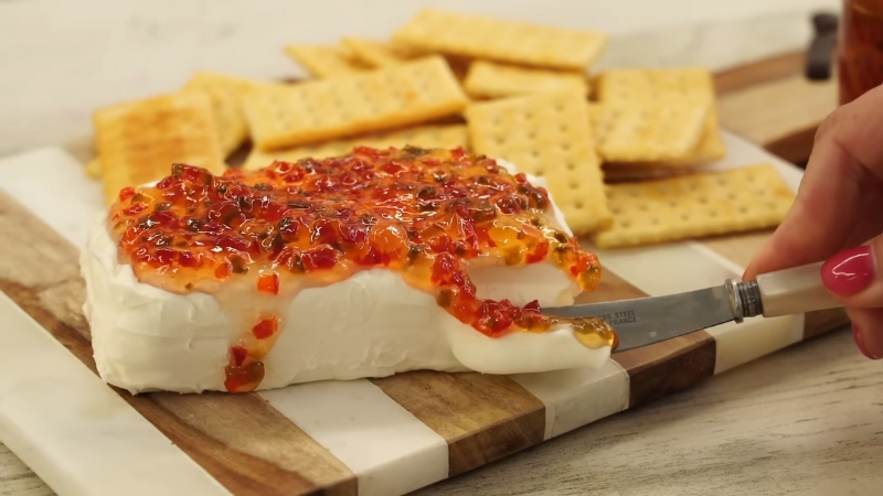 A block of cream cheese topped with homemade jalapeño jelly is being spread with a knife