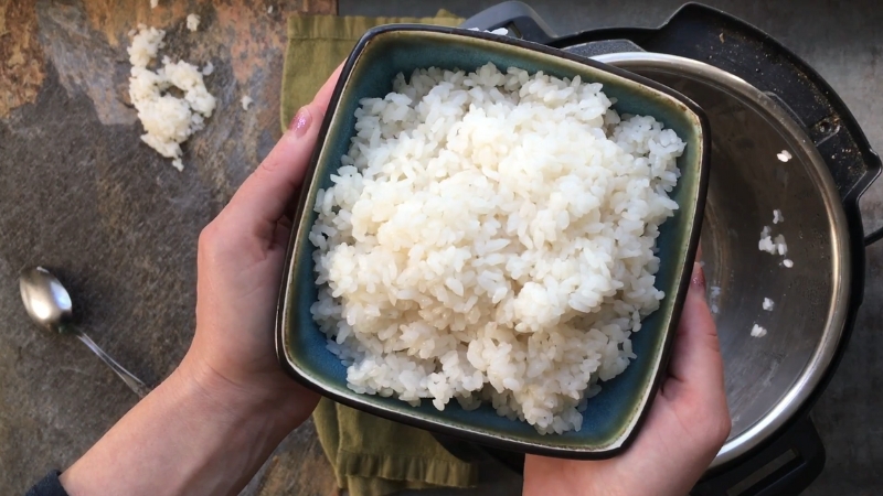 A bowl of freshly cooked and seasoned sushi rice is being held above an Instant Pot