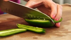A person slices fresh jalapeño peppers lengthwise on a wooden cutting board using a large kitchen knife