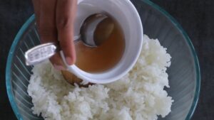 Sushi vinegar seasoning is being poured over freshly cooked rice in a glass bowl